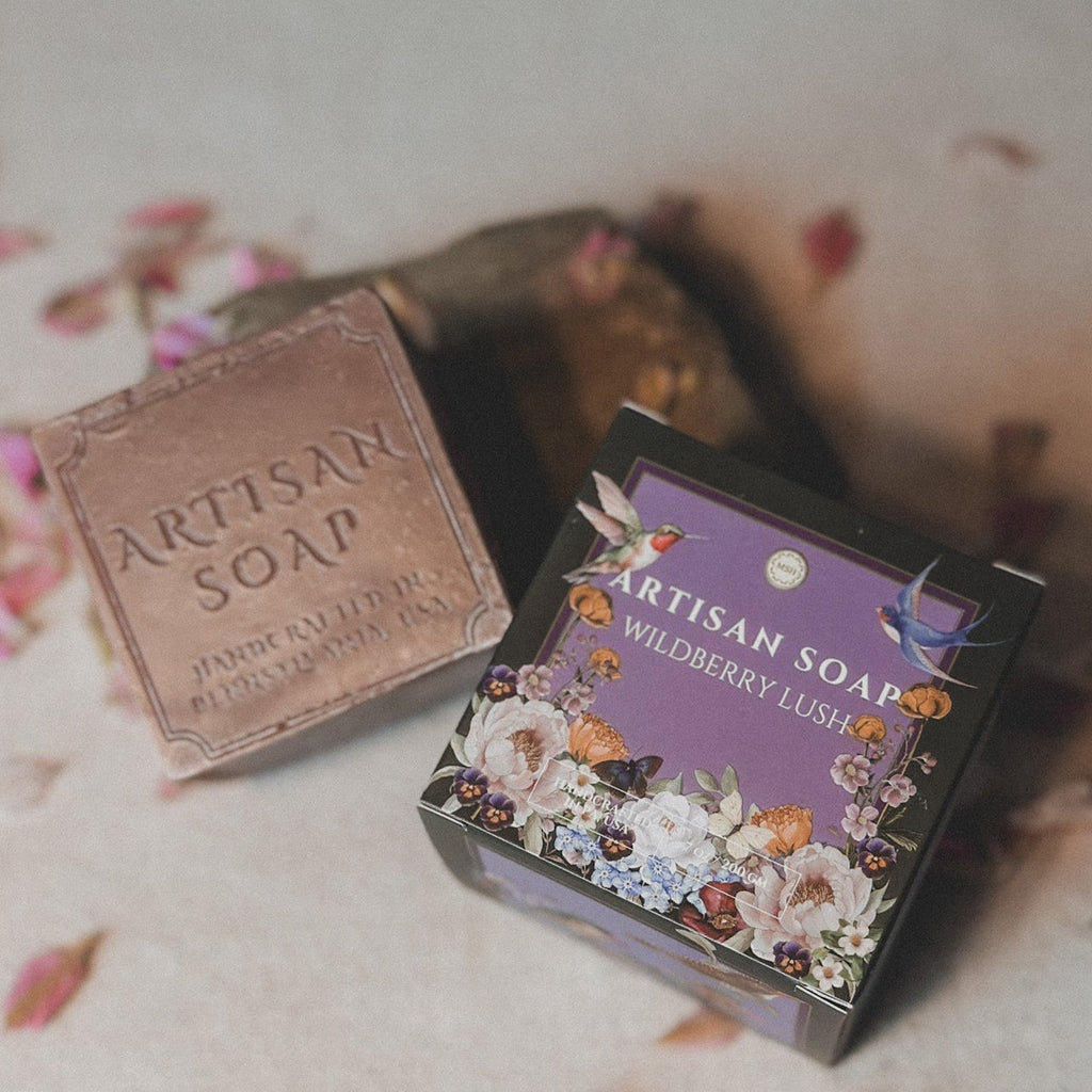 A bar of Artisan Soap labeled 'Wildberry Lush' placed next to its packaging which features floral designs. The bar has a textured, natural appearance and the packaging is adorned with an artistic depiction of berries and leaves.