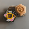 Hexagonal black box with floral design and bee emblem, next to a honeycomb-shaped product on a gray background