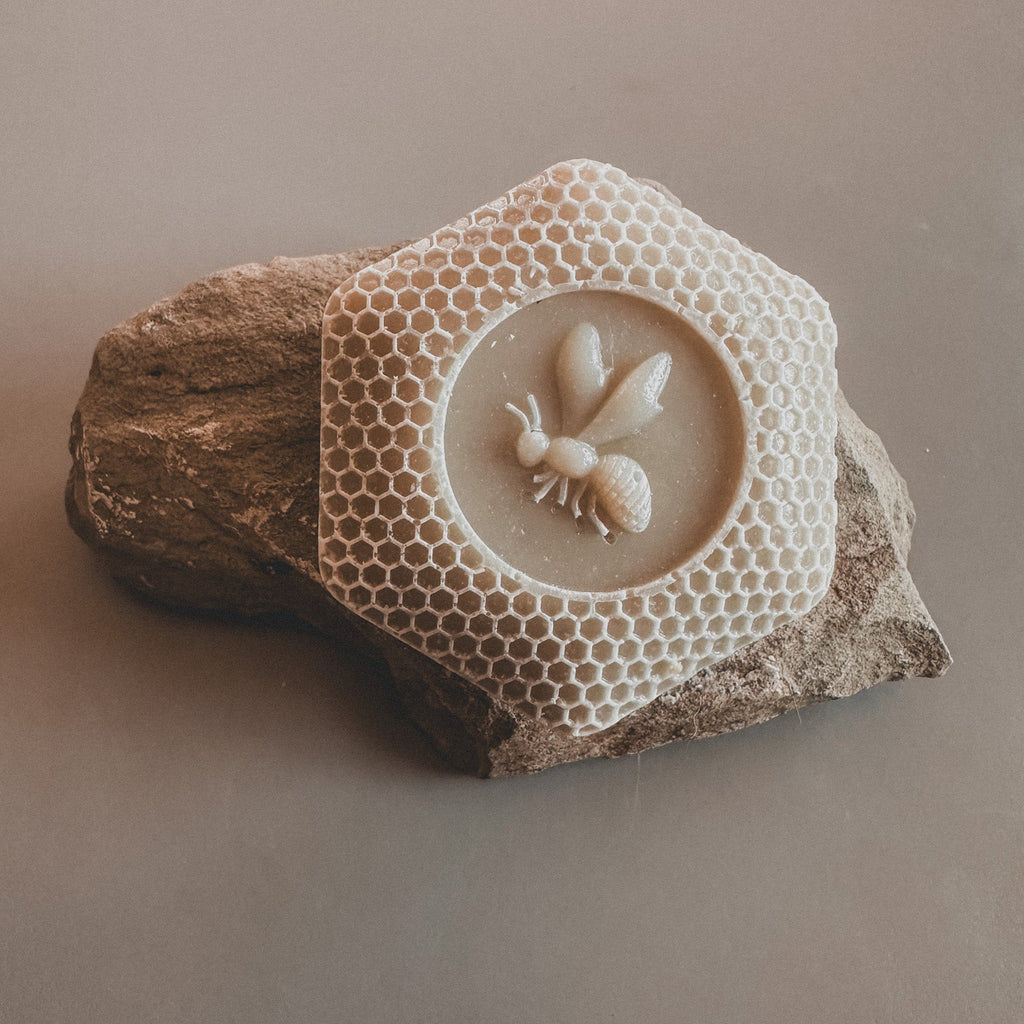A bar of soap designed to resemble a honeycomb with a bee motif in the center, placed on a natural looking rock.